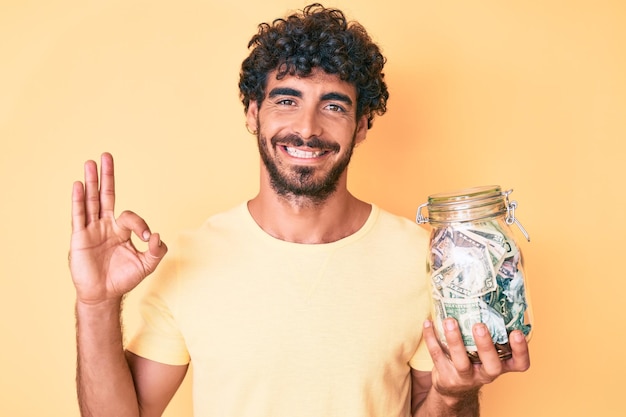 Beau jeune homme aux cheveux bouclés et ours tenant un pot avec des économies faisant signe ok avec les doigts, souriant amical gesticulant excellent symbole