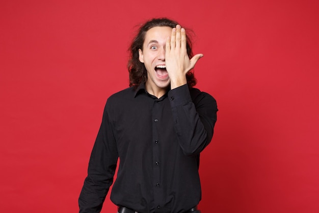 Beau jeune homme aux cheveux longs bouclé élégant en chemise noire posant isolé sur le portrait en studio de fond de mur rouge. Concept de mode de vie des émotions sincères des gens. Maquette de l'espace de copie. Couvrir les yeux avec la main.