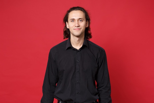 Beau Jeune Homme Aux Cheveux Longs Bouclé élégant En Chemise Noire Posant Isolé Sur Le Portrait En Studio De Fond De Mur Rouge. Concept De Mode De Vie Des émotions Sincères Des Gens. Maquette De L'espace De Copie. Regarder Le Sourire De La Caméra.