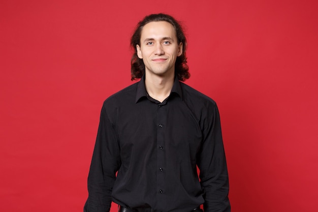 Beau jeune homme aux cheveux longs bouclé élégant en chemise noire posant isolé sur le portrait en studio de fond de mur rouge. Concept de mode de vie des émotions sincères des gens. Maquette de l'espace de copie. Regarder le sourire de la caméra.