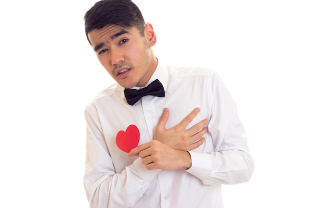 Beau jeune homme aux cheveux noirs en T-shirt blanc avec noeud papillon noir tenant un coeur en papier rouge