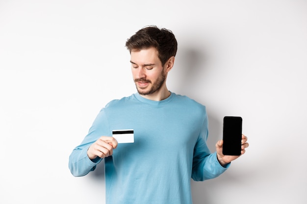 Beau Jeune Homme Avec Barbe, Montrant L'écran Du Smartphone Vide Et Regardant La Carte De Crédit En Plastique, Debout Sur Fond Blanc.