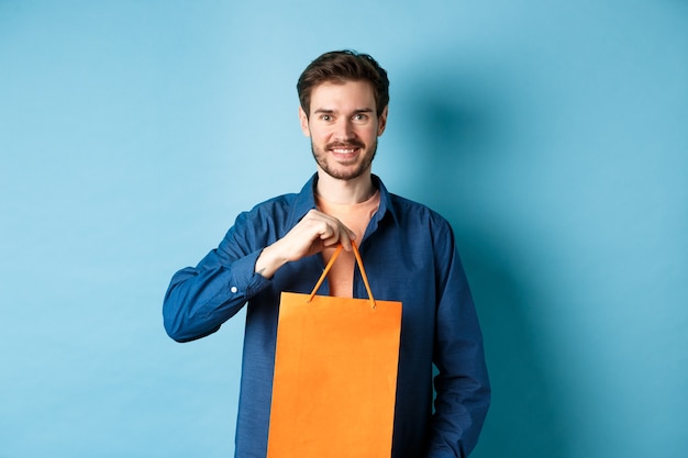 Beau jeune homme avec barbe, souriant et montrant le sac à provisions, achetant quelque chose en magasin, debout sur fond bleu.