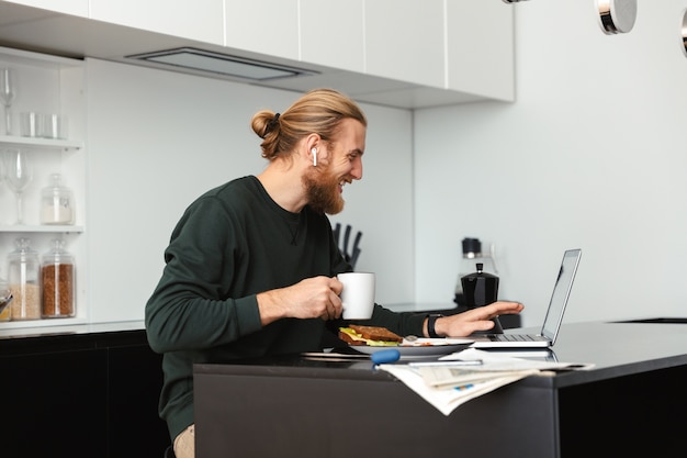 Beau jeune homme barbu assis à la cuisine, boire du café à l'aide d'un ordinateur portable.