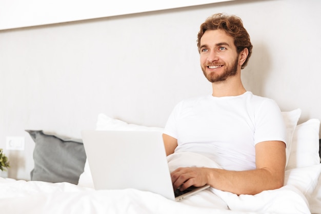 un beau jeune homme barbu au lit à la maison à l'aide d'un ordinateur portable.