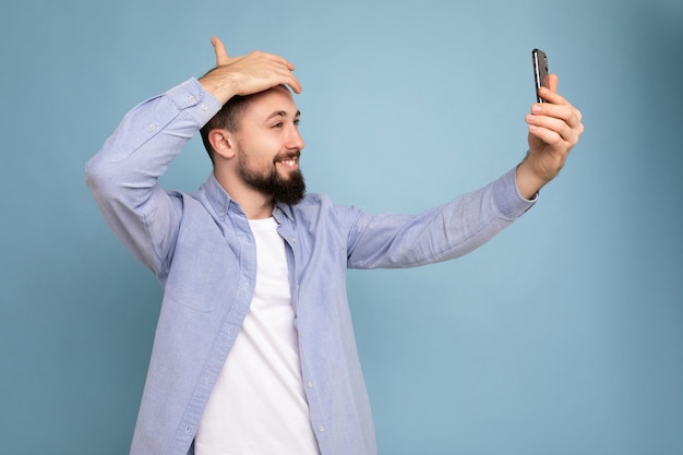 Beau jeune homme barbu brunet portant des vêtements élégants décontractés debout isolé sur un mur de mur bleu tenant un smartphone prenant selfie photo en regardant l'écran du téléphone mobile.