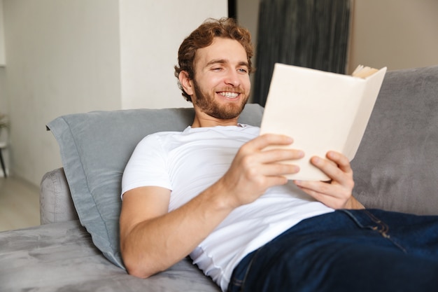 un beau jeune homme barbu sur un canapé à la maison, livre de lecture.