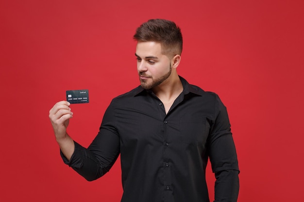 Beau jeune homme barbu en chemise noire classique posant isolé sur fond de mur rouge vif portrait en studio. Concept de mode de vie des émotions sincères des gens. Maquette de l'espace de copie. Détenir une carte bancaire de crédit.