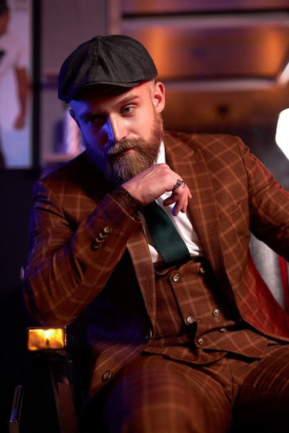 Beau jeune homme barbu dans un smoking assis dans une pièce sombre dans des vêtements à la mode