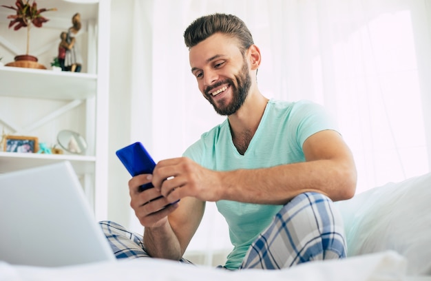 Beau jeune homme barbu dans des vêtements décontractés travaille à la maison assis sur le lit. Un mec confiant avec un ordinateur portable et un smartphone boit du café dans la chambre.