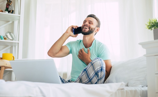 Photo beau jeune homme barbu dans des vêtements décontractés travaille à la maison assis sur le lit. un mec confiant avec un ordinateur portable et un smartphone boit du café dans la chambre.