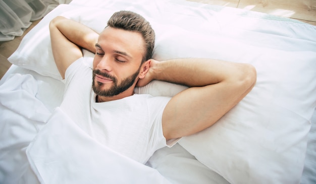 Beau jeune homme barbu dort dans un grand lit blanc dans une chambre confortable