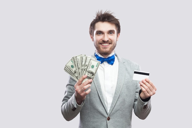 Beau jeune homme barbu heureux en costume gris et noeud papillon bleu debout et tenant une carte de crédit bancaire et de nombreux dollars en espèces avec un visage souriant à pleines dents. Intérieur, isolé, tourné en studio, fond gris