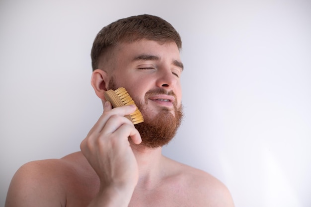 Beau jeune homme barbu Un homme brutal européen avec un poil de barbe peigne la barbe sur le visage avec une brosse à peigne Accessoire de produits de soins personnels pour hommes isolé sur fond blanc