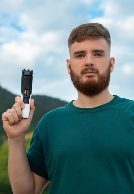 Beau jeune homme barbu Un homme brutal européen avec un poil de barbe se rase la barbe de coupe sur le visage avec une tondeuse Accessoire de produits d'auto-soins personnels masculins sur la nature Photo verticale