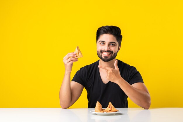 Beau jeune homme barbu indien mangeant des collations Samosa alors qu'il était assis à une table blanche sur fond jaune