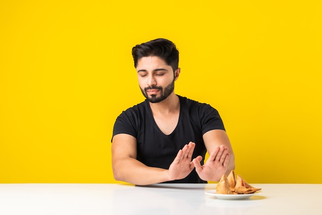 Beau jeune homme barbu indien mangeant des collations Samosa alors qu'il était assis à une table blanche sur fond jaune