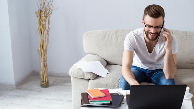 Beau jeune homme barbu souriant assis sur un canapé à la maison travaillant sur son ordinateur portable et son téléphone