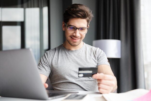 Beau jeune homme barbu souriant portant des vêtements décontractés à l'aide d'un ordinateur portable assis à la table à la maison, montrant une carte de crédit en plastique, faisant du shopping