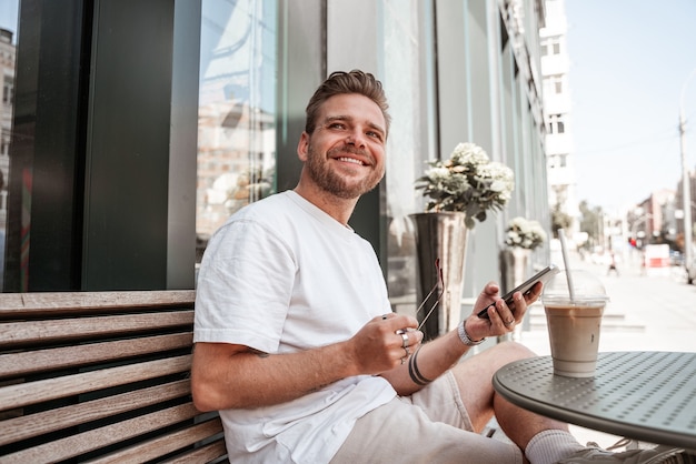 Beau jeune homme blond assis sur la terrasse du café avec une boisson au café souriant avec un smartphone mobile sur fond de réflexion du bâtiment en verre de la rue de la grande ville. Hipster millénaire le jour ensoleillé d'été