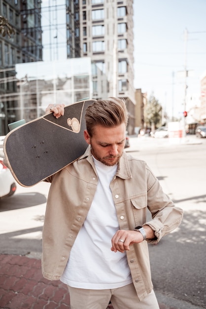 Beau jeune homme blond skateboarder tenant un longboard sur l'épaule à la sérieuse montre-bracelet à la hâte sur fond de construction urbaine. Porte une tenue décontractée en denim beige. Concept de loisirs extérieur.