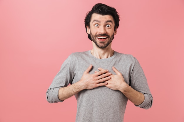 Beau jeune homme brune barbu choqué portant chandail debout sur rose, célébrant