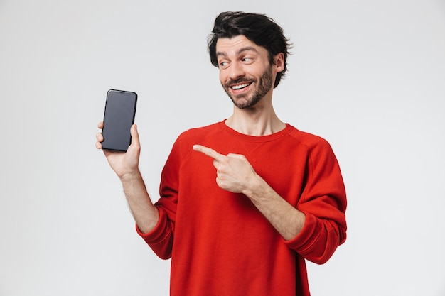 Beau jeune homme brune barbu excité portant chandail debout sur blanc, montrant un téléphone mobile