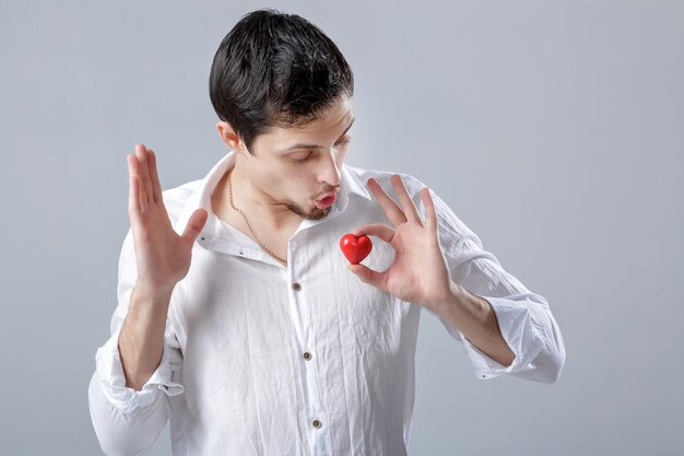 Beau jeune homme brune en chemise blanche tient en mains un coeur rouge sur fond gris.