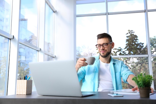 Beau jeune homme buvant du café tout en travaillant avec un ordinateur portable à la maison