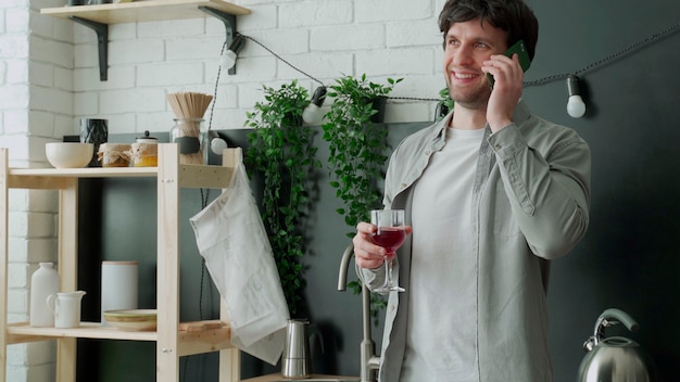 Beau jeune homme buvant du vin et parlant au téléphone alors qu'il était assis dans la cuisine à la maison