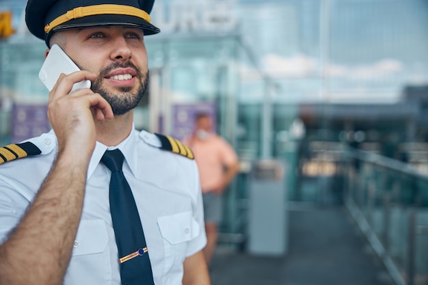 Beau jeune homme capitaine de compagnie aérienne ayant une conversation téléphonique et souriant à l'extérieur