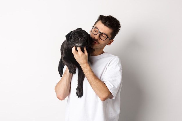 Beau jeune homme caresse son mignon chien noir, grattant le carlin tout en tenant l'animal sur l'épaule, debout sur fond blanc.