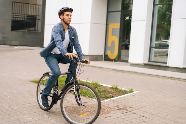 Un beau jeune homme caucasien portant un casque de protection sort pour une balade à vélo dans les rues de la ville sur fond flou de la porte d'entrée de l'immeuble. Un cycliste fait du vélo à l'extérieur en milieu urbain.