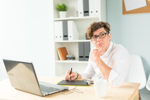 Beau jeune homme en chemise blanche à l'aide de tablette graphique à table en bois