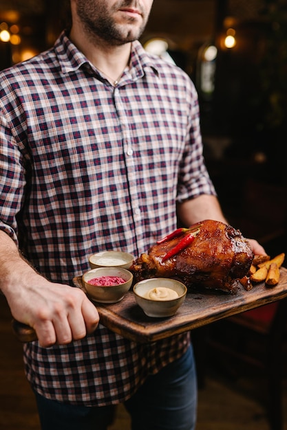 Beau jeune homme en chemise à carreaux tenant un plateau en bois avec jarret de porc dans un pub