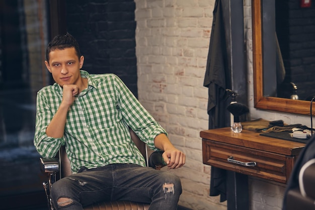 Beau jeune homme en chemise et jeans en regardant la caméra et assis sur une chaise au salon de coiffure. Il a l'air confiant