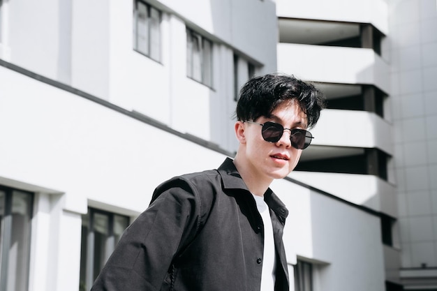 Beau jeune homme en chemise noire debout sur fond de bâtiments de la ville et regardant à travers des lunettes de soleil noires