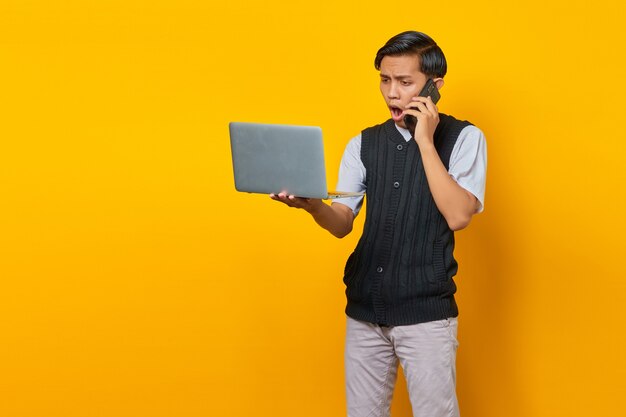 Un beau jeune homme choqué parlant sur un smartphone et regardant un écran d'ordinateur portable sur fond jaune