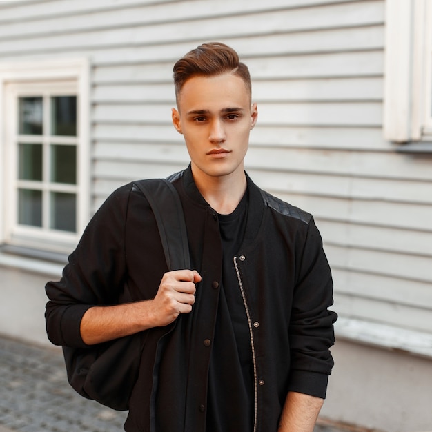 Beau Jeune Homme Avec Une Coiffure Dans Une Veste Avec Un Sac à Dos Près D'une Maison En Bois