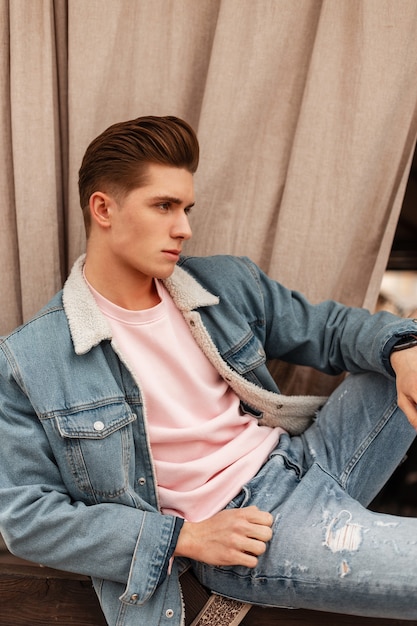 Beau jeune homme avec une coiffure à la mode dans des vêtements de jeans pour jeunes bleus à la mode de la nouvelle collection d'été reste dans la rue à l'extérieur. Beau modèle de mec élégant dans des vêtements décontractés en denim de mode en ville.