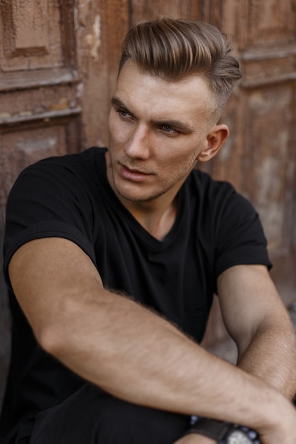 Beau jeune homme avec une coiffure en t-shirt noir assis près d'une vieille porte vintage