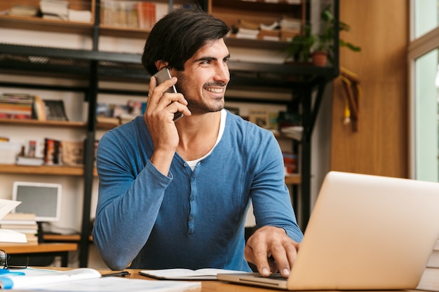 Beau jeune homme confiant assis au bureau de la bibliothèque, travaillant / étudiant, utilisant un ordinateur portable, parler au téléphone mobile