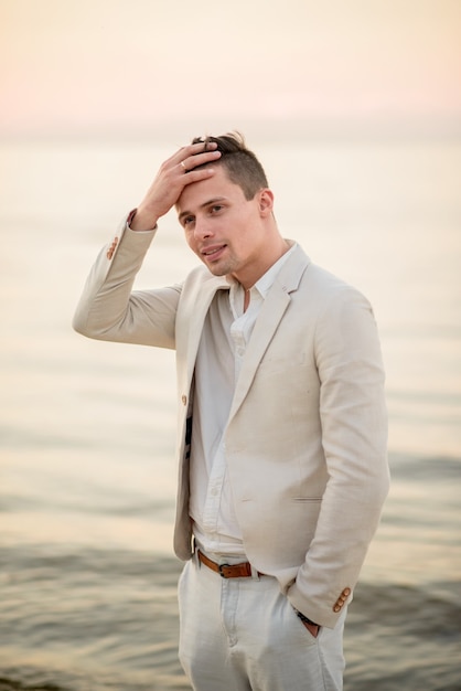 Beau jeune homme en costume blanc au bord de la mer au coucher du soleil