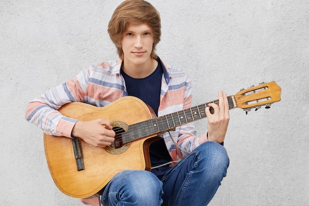 Beau jeune homme avec coupe de cheveux élégante portant chemise et jeans jouant de la guitare, chantant des chansons