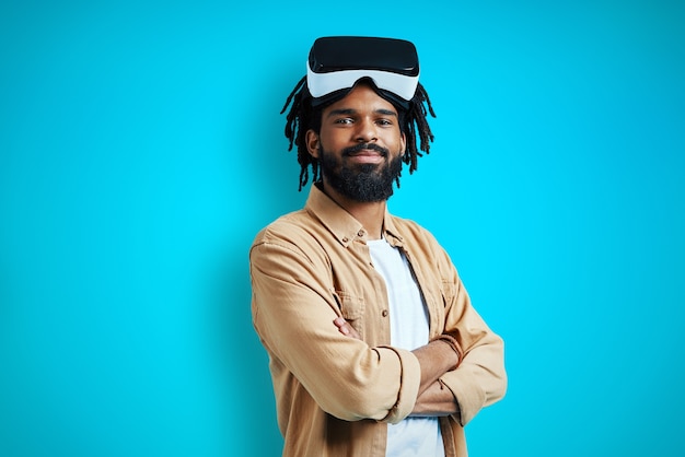 Beau jeune homme dans des lunettes de réalité virtuelle regardant la caméra et souriant en se tenant debout contre le mur bleu