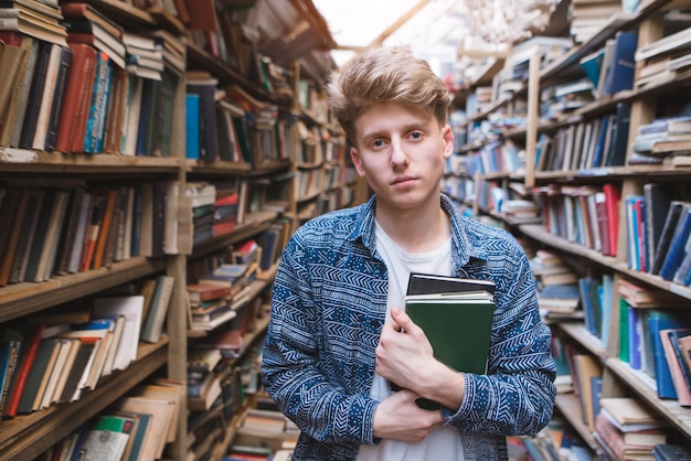 Beau jeune homme debout dans une bibliothèque publique avec des livres dans ses mains et regardant la caméra