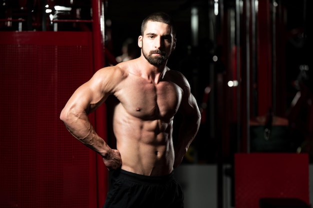 Beau jeune homme debout fort dans la salle de gym et muscles fléchissants - Modèle de forme physique de bodybuilder athlétique musclé posant après des exercices