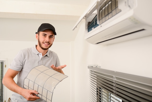 Beau jeune homme électricien nettoyant le filtre à air sur une unité intérieure de système de climatisation dans une maison client