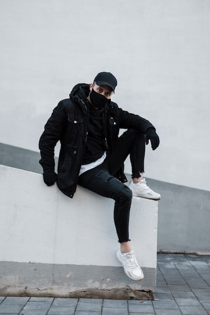Beau jeune homme élégant avec une casquette noire et un masque médical de protection dans une veste noire, un sweat à capuche, un jean et des baskets se trouve près du mur blanc dans la rue