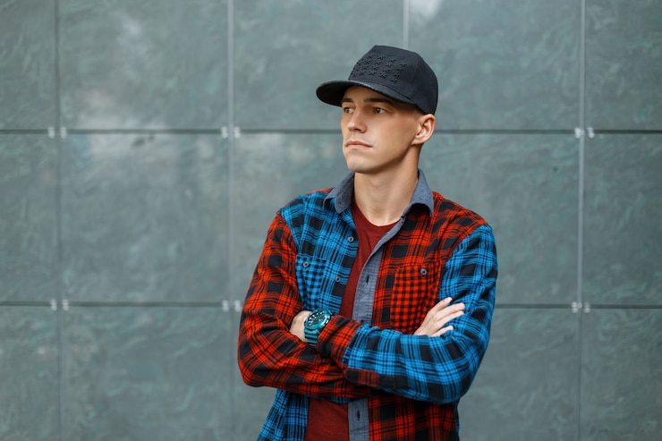 Photo beau jeune homme élégant dans un t-shirt élégant dans une chemise à carreaux bleue et rouge à la mode dans une casquette de basket-ball à la mode noire se tient près d'un mur gris vintage.